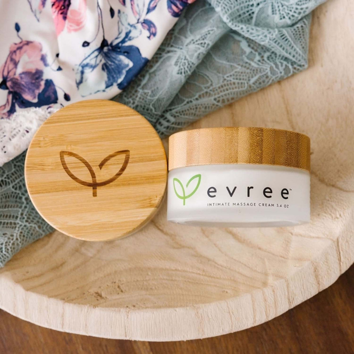 Open jar of evree intimate massage cream beside its bamboo lid, on a wooden board with silk and lace